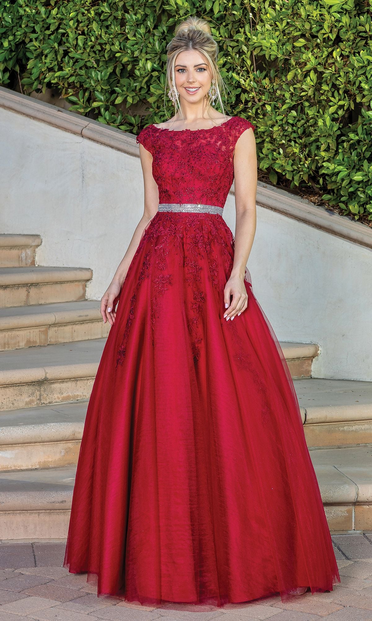 A woman stands outdoors on stone steps, framed by lush greenery, wearing the Corset-Back Illusion-Sweetheart Long Ball Gown featuring rich red embroidered lace, a silver belt, and elegant detailing.